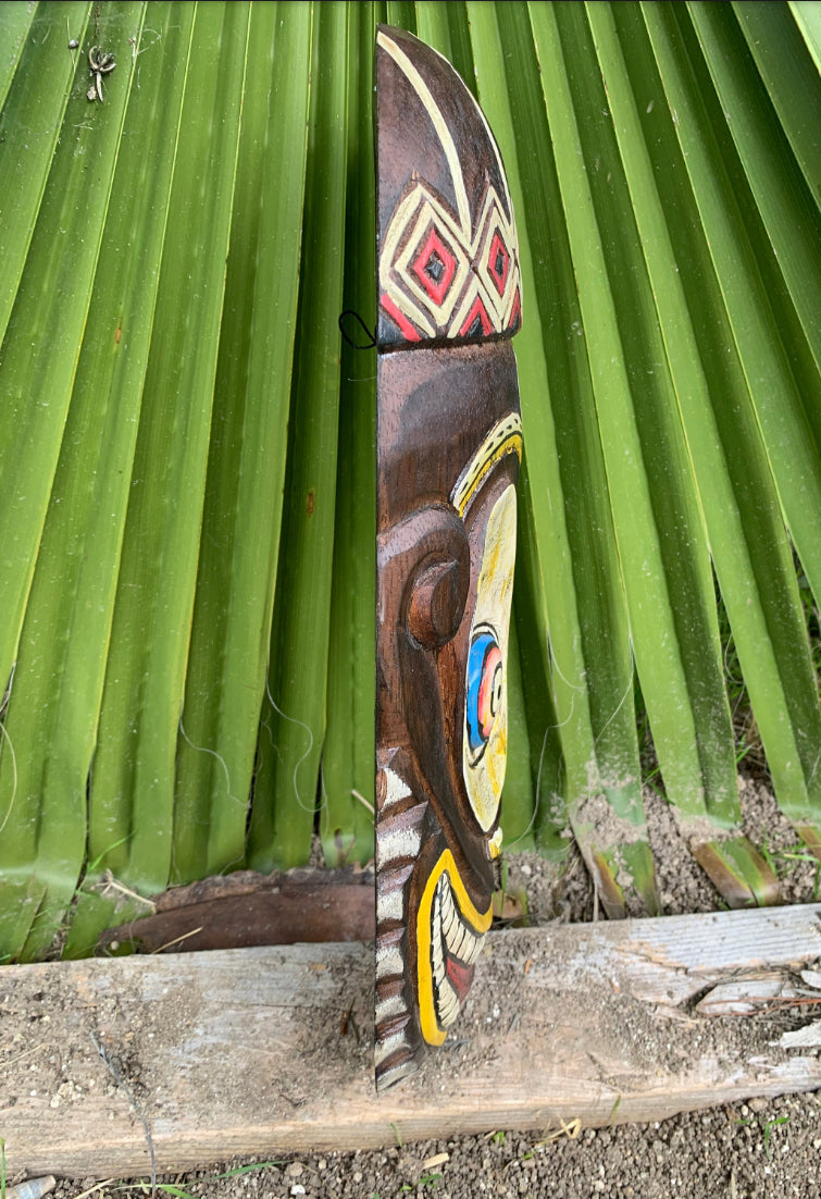 Colorful wooden tiki mask carved with geometric patterns leaning against green palm leaves.